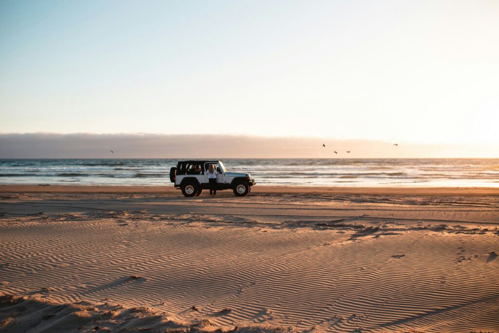 A 4x4 vehicle parked on a sandy beach at sunset, with a person standing beside it and the ocean in the background.