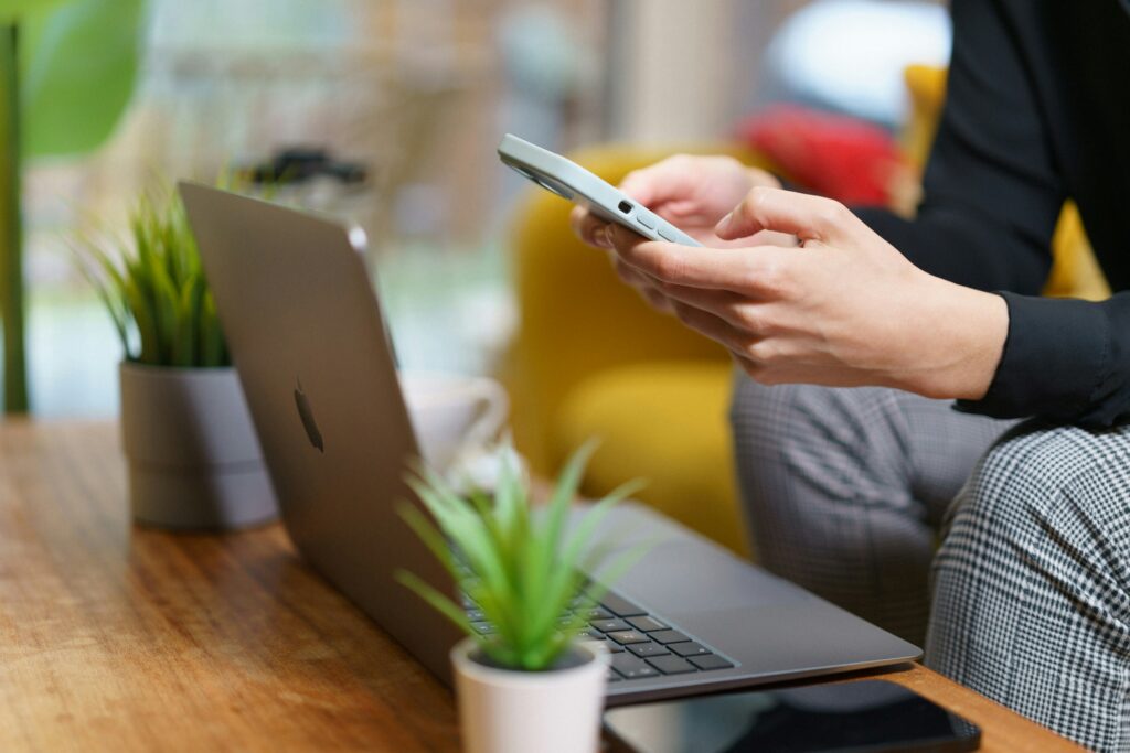 Person using a smartphone to buy tyres online while sitting at a desk with a laptop and small potted plants